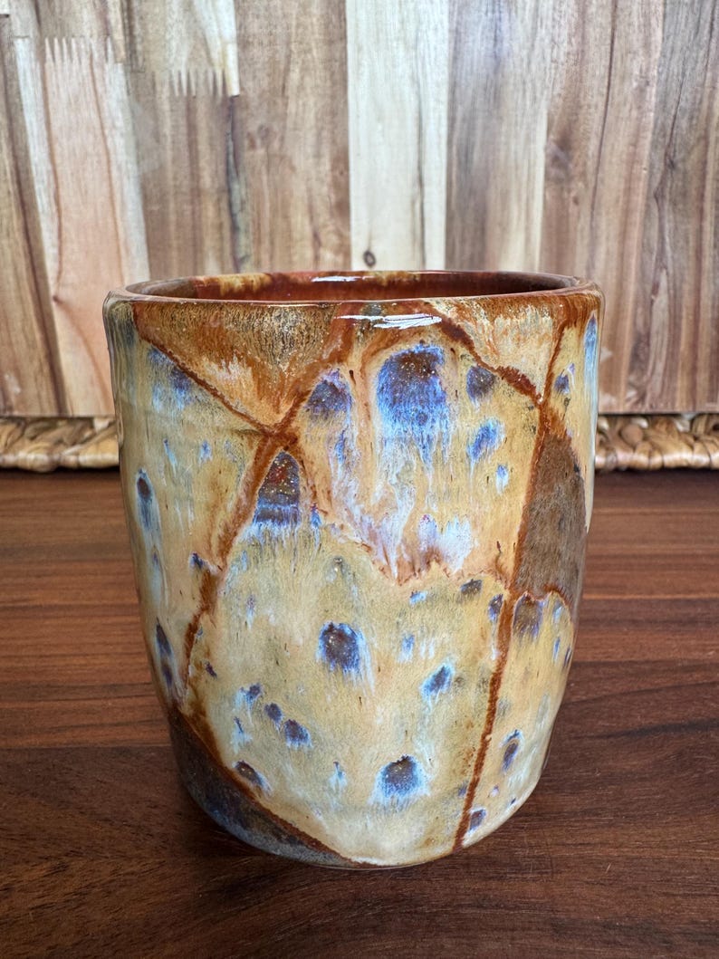 May include: A cylindrical ceramic tumbler featuring a glossy glaze with a mottled pattern of tan, brown, and deep blue hues. The surface displays a network of dark brown lines that create a segmented, crackled appearance across the exterior.