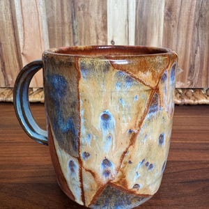 May include: A ceramic mug featuring a glossy glaze with a marbled pattern in shades of tan, brown, and blue. The mug has a cylindrical body and a curved handle on the left side, showcasing a smooth, reflective finish with distinct color transitions.