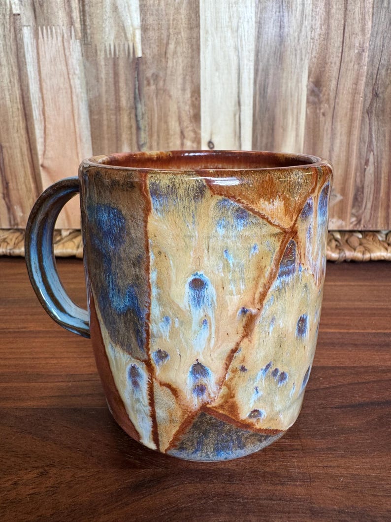 May include: A ceramic mug featuring a glossy glaze with a marbled pattern in shades of tan, brown, and blue. The mug has a cylindrical body and a curved handle on the left side, showcasing a smooth, reflective finish with distinct colour transitions.