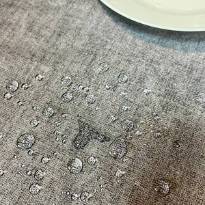 May include: A close-up of a gray fabric surface with water droplets scattered across it. The droplets are round and clear, and they are reflecting the light. A white plate is visible in the background.