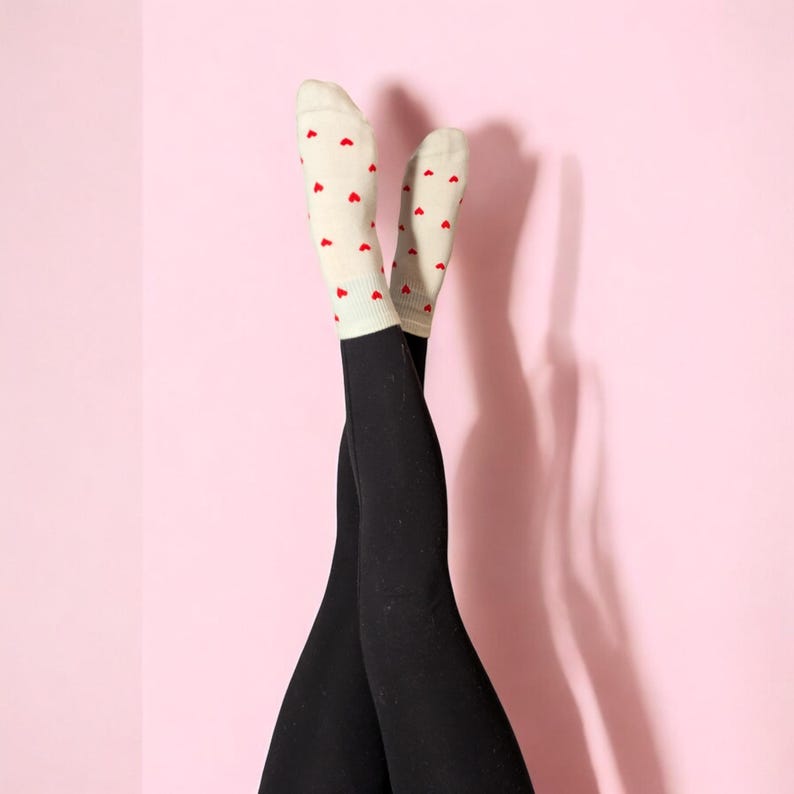 May include: A pair of black leggings with white socks that have red hearts printed on them. The socks are pulled up to the knees and the legs are crossed at the ankles.