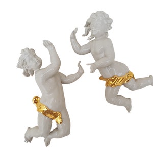 Könnte beinhalten: Zwei weiße Porzellan-Puttenfiguren mit goldenen Akzenten. Einer der Putten greift nach oben, während der andere sich in einer spielerischen Pose befindet. Die Putten sind mit lockigem Haar und goldfarbenen Schärpen detailliert. Die Figuren sind vor weißem Hintergrund.