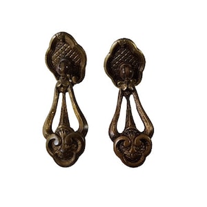 Peut inclure: Deux poignées de cabinet en laiton ornées d'un motif décoratif. Les poignées ont une forme de larme avec un motif floral en bas et un motif carré en haut.