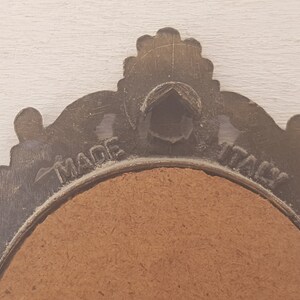 May include: A close-up of the back of a round, ornate, metal picture frame with a brown cardboard backing. The frame is inscribed with the words "MADE IN ITALY".