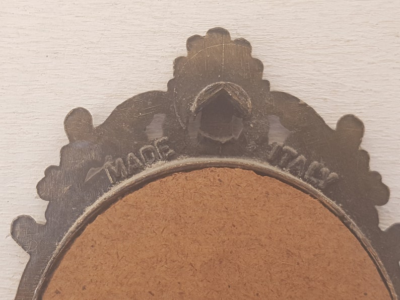 May include: A close-up of the back of a round, ornate, metal picture frame with a brown cardboard backing. The frame is inscribed with the words "MADE IN ITALY".