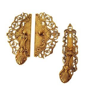May include: Ornate, antique-style brass door hardware set. The set includes two half-circle backplates and a long, decorative pull. The hardware features intricate scrollwork and a detailed, textured surface.