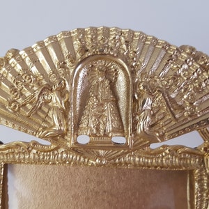 May include: Gold-toned picture frame with ornate details. The top of the frame features a fan-shaped design with a central image of a religious figure. Two angels are flanking the central image.