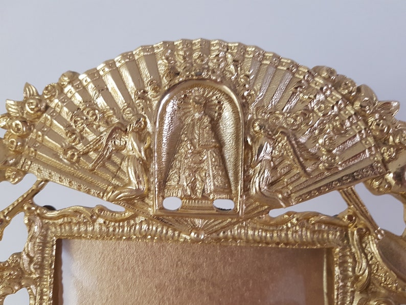 May include: Gold-toned picture frame with ornate details. The top of the frame features a fan-shaped design with a central image of a religious figure. Two angels are flanking the central image.