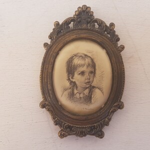 May include: A framed drawing of a child with short brown hair, wearing a light-colored shirt, in a gold oval frame with ornate details.
