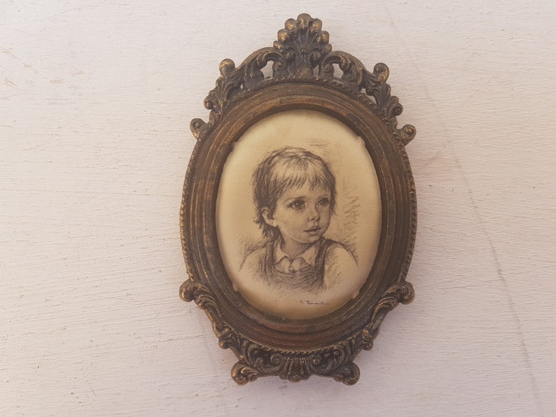 May include: A framed drawing of a child with short brown hair, wearing a light-colored shirt, in a gold oval frame with ornate details.