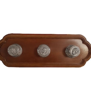 Pode incluir: Um cabide de parede de madeira com três ganchos decorativos. Cada gancho é uma peça redonda de metal prateado com um design em relevo.