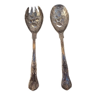 May include: A set of two silver serving utensils with ornate handles. The utensils have a face design with holes for serving food.