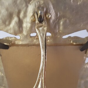 May include: Gold-toned metal picture frame with an ornate design. The frame has a stand on the back and two small metal tabs on the sides.