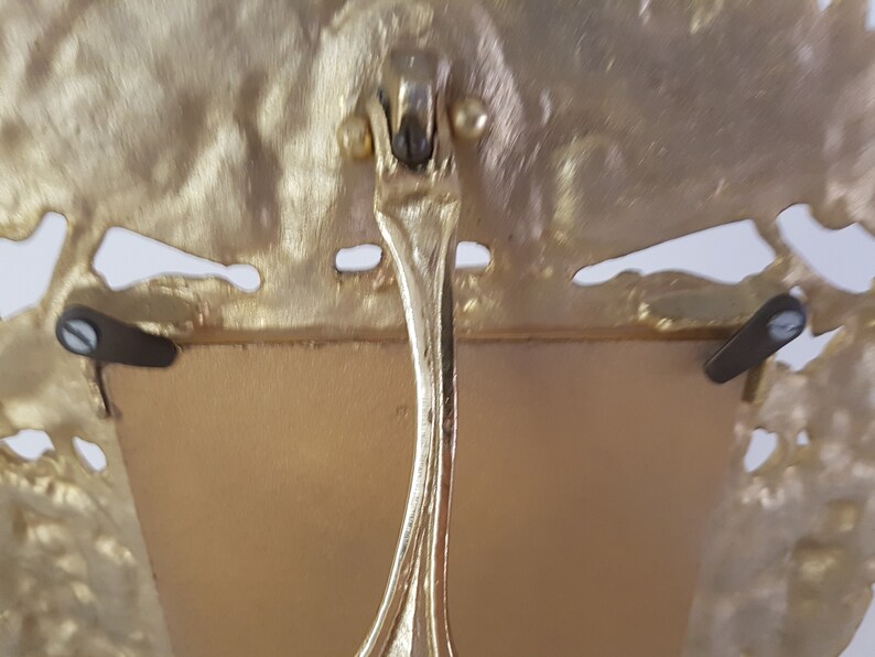 May include: Gold-toned metal picture frame with an ornate design. The frame has a stand on the back and two small metal tabs on the sides.