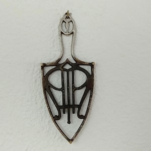 May include: A black metal trivet in the shape of a shield with a decorative design. The trivet has a handle on top.