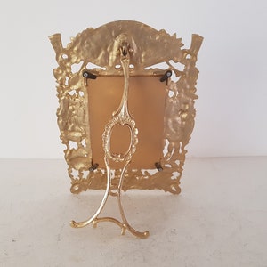 May include: A gold-toned metal picture frame with an ornate design. The frame has a stand on the back and a rectangular opening for a picture.