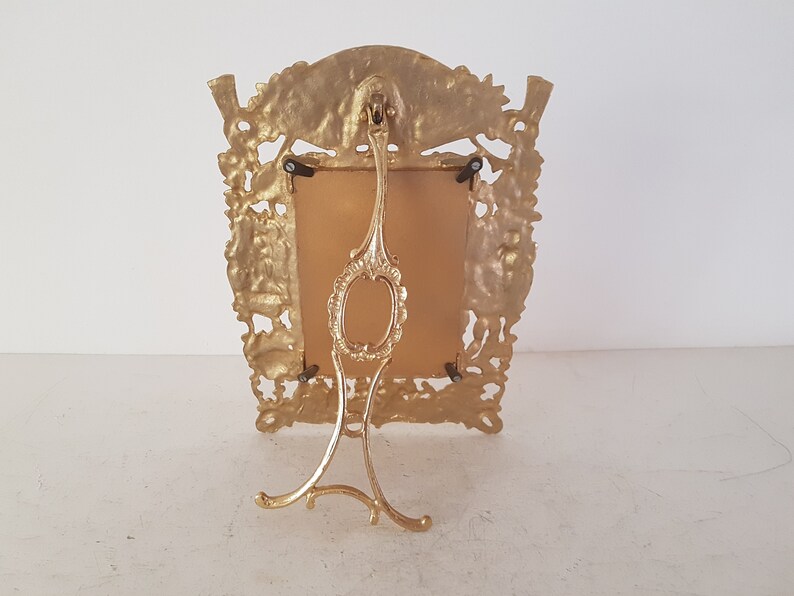May include: A gold-toned metal picture frame with an ornate design. The frame has a stand on the back and a rectangular opening for a picture.