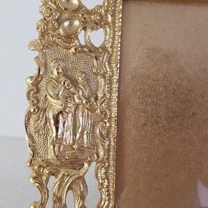 May include: A gold-colored picture frame with an ornate design featuring two figures in relief. The frame has a rectangular shape and a smooth, reflective surface.