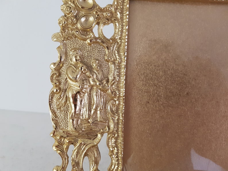May include: A gold-colored picture frame with an ornate design featuring two figures in relief. The frame has a rectangular shape and a smooth, reflective surface.