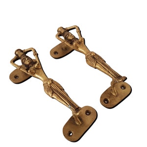 May include: A pair of gold-toned door handles shaped like a woman holding a pitcher on her head. The handles are attached to a rectangular base with two holes for mounting.