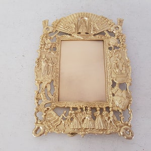 May include: A gold-toned metal picture frame with ornate details and a raised design. The frame features a central rectangular opening for a photo and is decorated with figures and floral motifs.