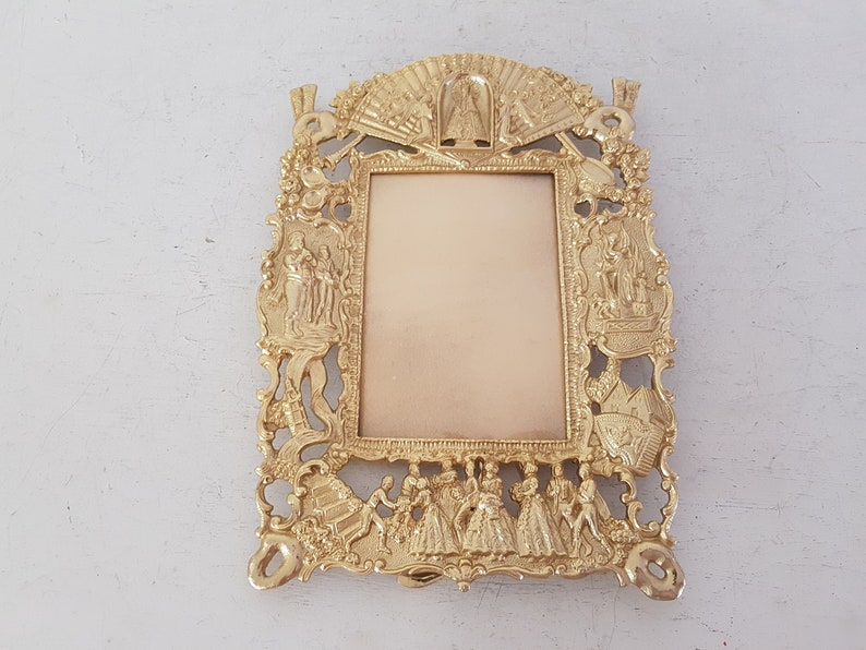 May include: A gold-toned metal picture frame with ornate details and a raised design. The frame features a central rectangular opening for a photo and is decorated with figures and floral motifs.