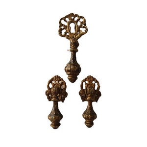May include: Three ornate gold-toned metal drawer pulls with intricate designs. The top pull has a keyhole design, while the two bottom pulls have a more floral design.