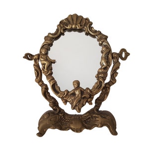 May include: A gold-toned, ornate tabletop mirror with a decorative cherub design. The mirror has an oval-shaped glass and a stand with three legs.