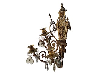 May include: A gold-toned metal wall sconce with three candle holders and crystal drops. The sconce has an ornate design with intricate details.