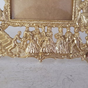 May include: A gold-toned metal picture frame with an ornate design. The frame features a raised design of people in a procession, with intricate details and a textured surface.