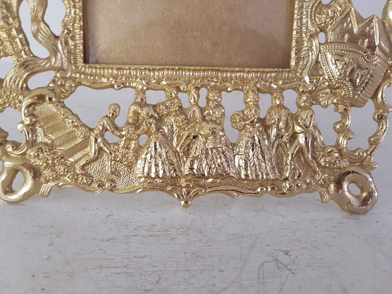 May include: A gold-toned metal picture frame with an ornate design. The frame features a raised design of people in a procession, with intricate details and a textured surface.