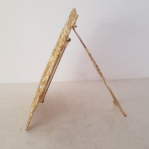 May include: A gold-colored metal picture frame with an ornate design. The frame has a stand on the back, allowing it to be displayed on a table or shelf.