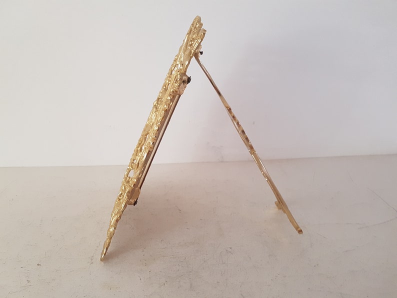 May include: A gold-colored metal picture frame with an ornate design. The frame has a stand on the back, allowing it to be displayed on a table or shelf.