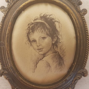May include: A framed drawing of a young girl with brown hair and eyes, wearing a light-colored dress. The frame is oval-shaped and made of gold-toned metal.