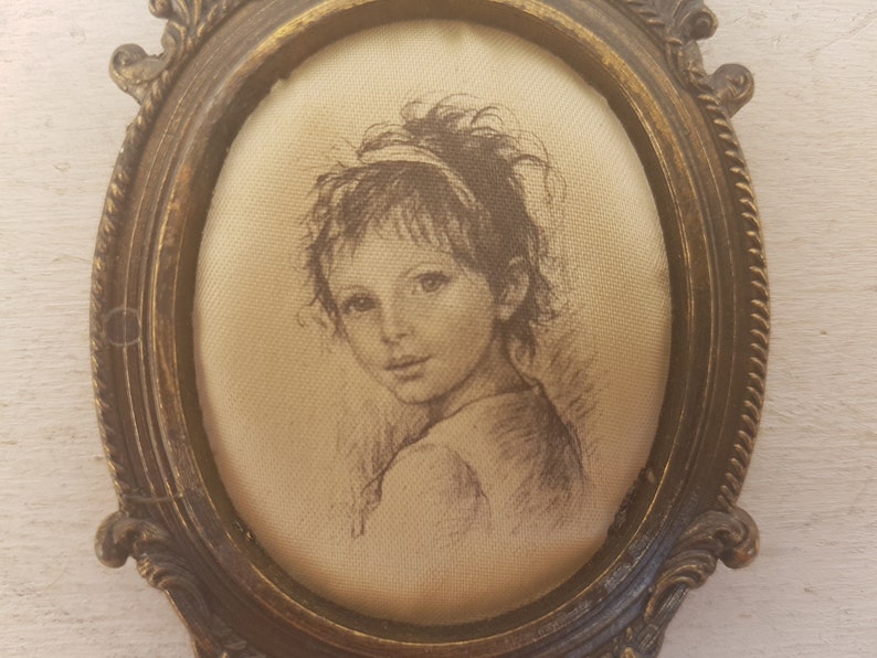 May include: A framed drawing of a young girl with brown hair and eyes, wearing a light-colored dress. The frame is oval-shaped and made of gold-toned metal.