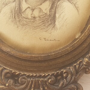 May include: A framed drawing of a person in a gold-colored ornate frame. The drawing is on a cream-colored fabric and is signed "F. Vannetten".