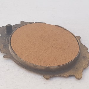 May include: A small, ornate, gold-toned metal picture frame with a brown cardboard backing. The frame has a decorative, floral design.