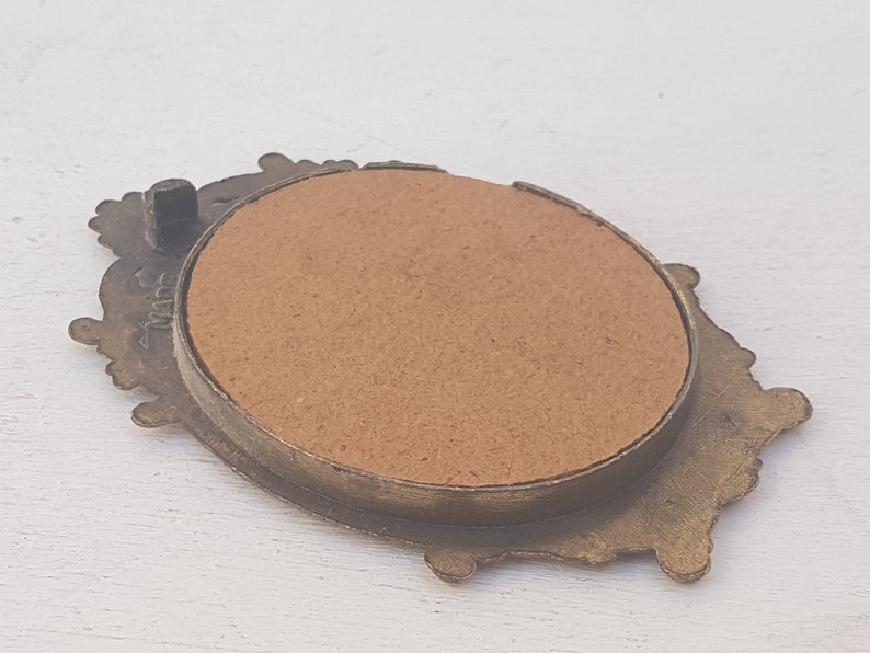 May include: A small, ornate, gold-toned metal picture frame with a brown cardboard backing. The frame has a decorative, floral design.