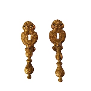 Könnte beinhalten: Zwei verzierte, goldfarbene Schlüssellochschilder. Jedes hat eine dekorative Oberseite mit einem Schlüsselloch, einen Mittelteil und einen tropfenförmigen Boden. Das Design umfasst florale und schnörkelartige Details.