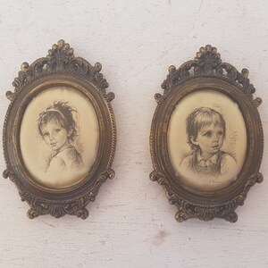 May include: Two oval framed portraits of children. The frames are ornate and gold in color. The portraits are in black and white and depict a girl and a boy.