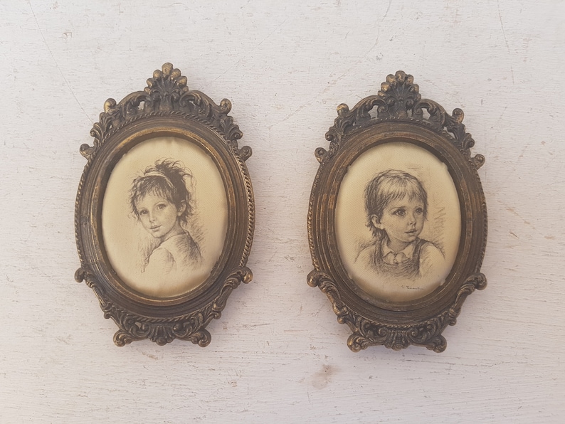 May include: Two oval framed portraits of children. The frames are ornate and gold in color. The portraits are in black and white and depict a girl and a boy.