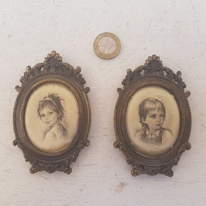 May include: Two oval-shaped framed portraits of children. The frames are ornate and gold-colored. The portraits are in black and white and depict a girl and a boy.