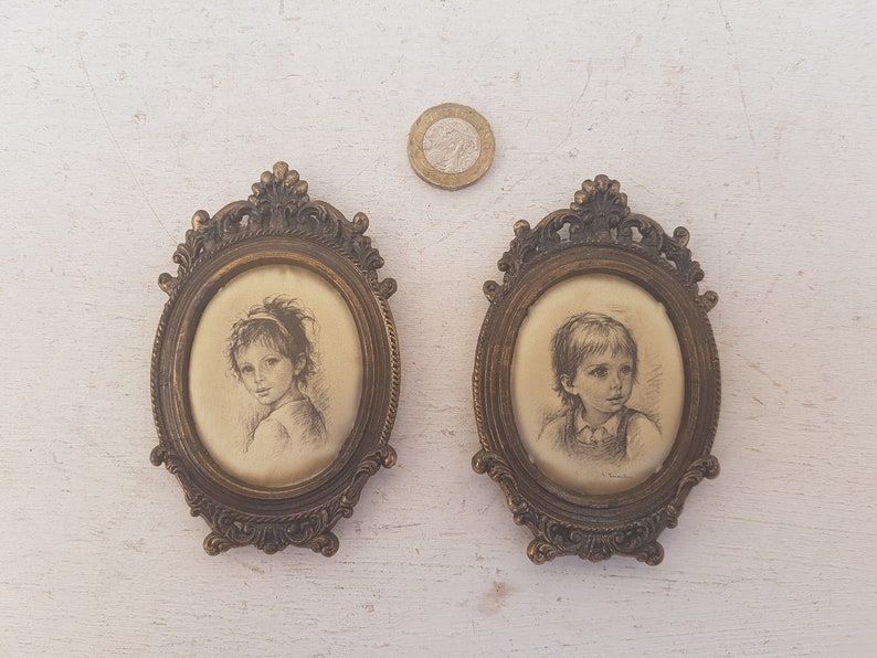 May include: Two oval-shaped framed portraits of children. The frames are ornate and gold-colored. The portraits are in black and white and depict a girl and a boy.
