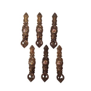 May include: Seven ornate, bronze-colored wall hooks. Each hook features a decorative design with a small, circular image in the center. The hooks have a detailed, vintage aesthetic, with a scrolled base and top.