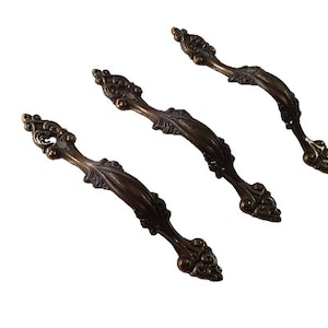 Peut inclure: Trois poignées de porte d'armoire de style antique ornées, avec une finition bronze foncé. Chaque poignée présente un motif décoratif enroulé.