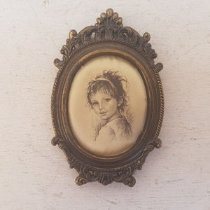 May include: A vintage oval-shaped picture frame with an ornate gold-toned metal design. The frame holds a black and white drawing of a young girl with a headband.