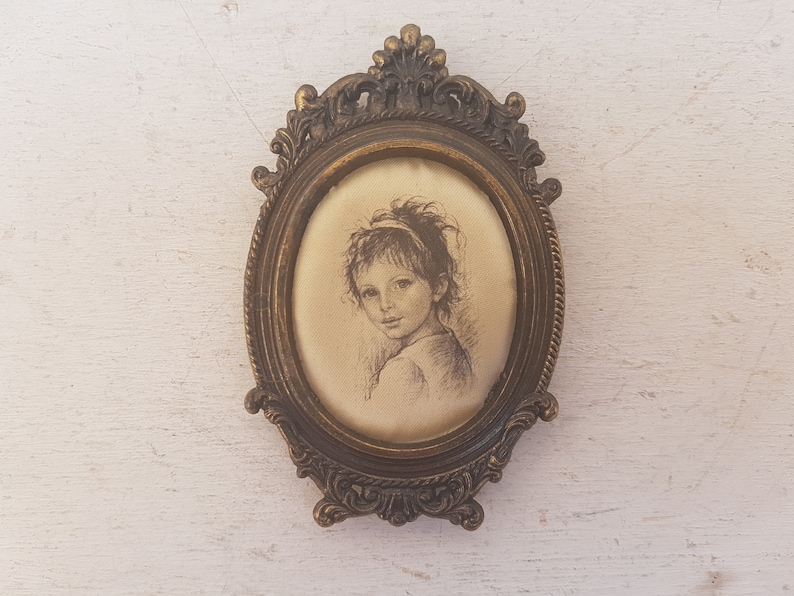 May include: A vintage oval-shaped picture frame with an ornate gold-toned metal design. The frame holds a black and white drawing of a young girl with a headband.