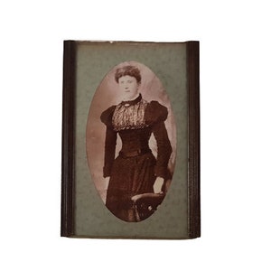 May include: A black and white portrait of a woman in a dark dress with a lace collar. The portrait is framed in a dark wood frame with a light green mat.