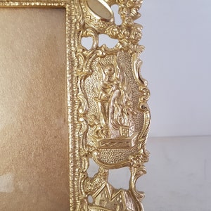 May include: A gold-colored picture frame with an ornate design. The frame has a raised, detailed design of a figure holding a staff, standing on a platform. The frame is made of metal.