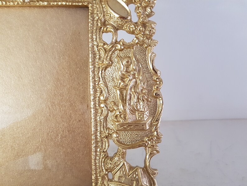 May include: A gold-colored picture frame with an ornate design. The frame has a raised, detailed design of a figure holding a staff, standing on a platform. The frame is made of metal.